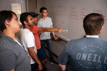 five people gather in discussion at a whiteboard